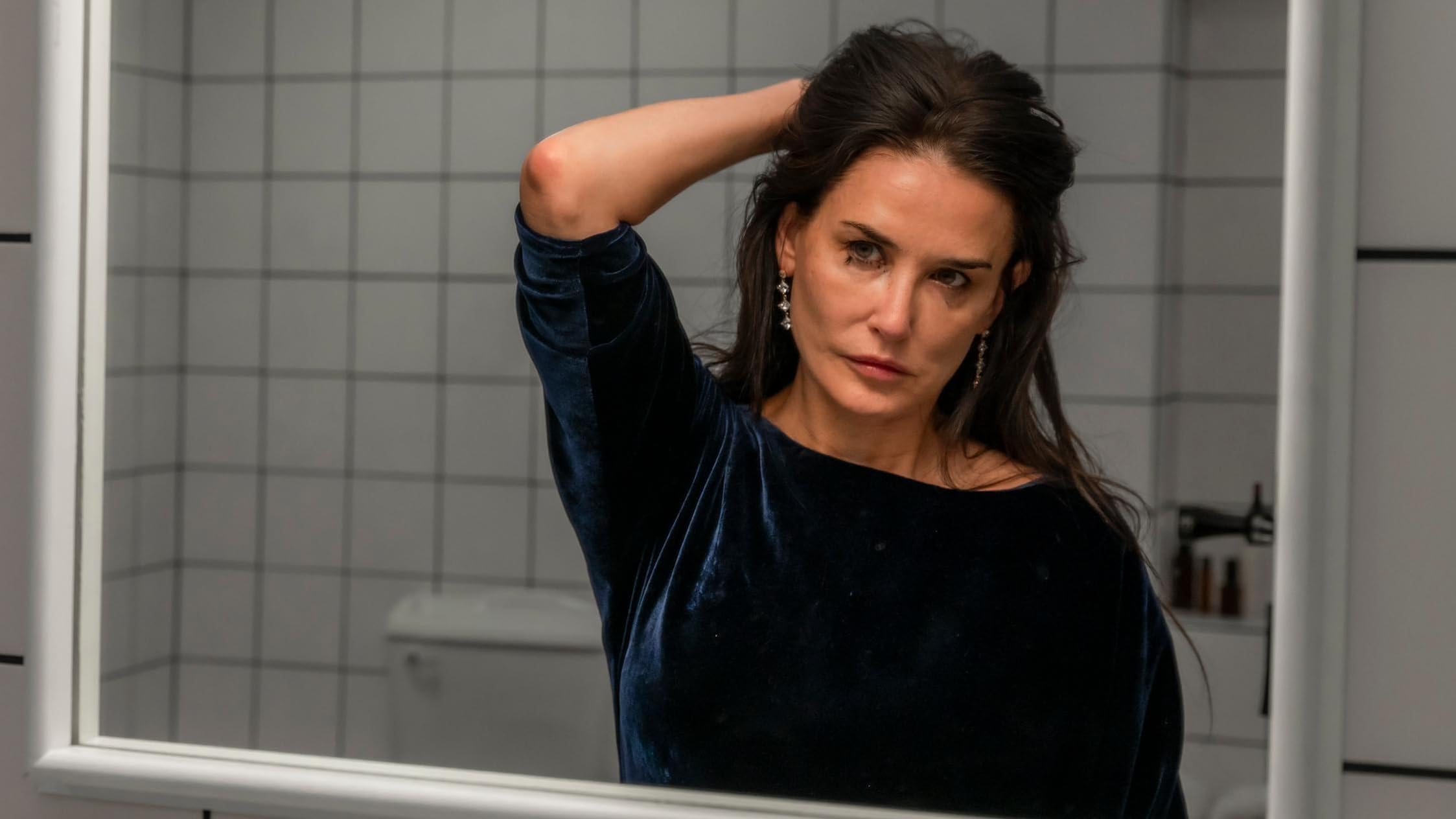El personaje de Demi Moore sentada en una gran bañera, con expresión contemplativa y vulnerable, rodeada de un baño lujoso pero estéril con superficies de mármol e iluminación suave, evocando una sensación de soledad e introspección