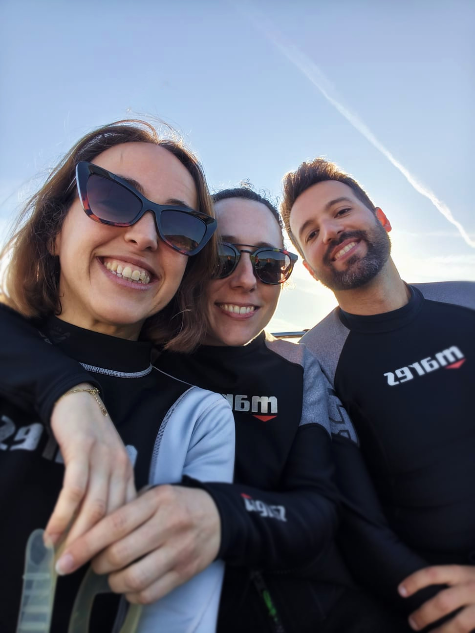 Un grupo de buceadores equipados con trajes de neopreno y botellas de oxígeno caminando por un muelle de madera en dirección al punto de inmersión, rodeados de aguas tranquilas y un paisaje costero sereno, transmitiendo una sensación de anticipación y camaradería antes de sumergirse